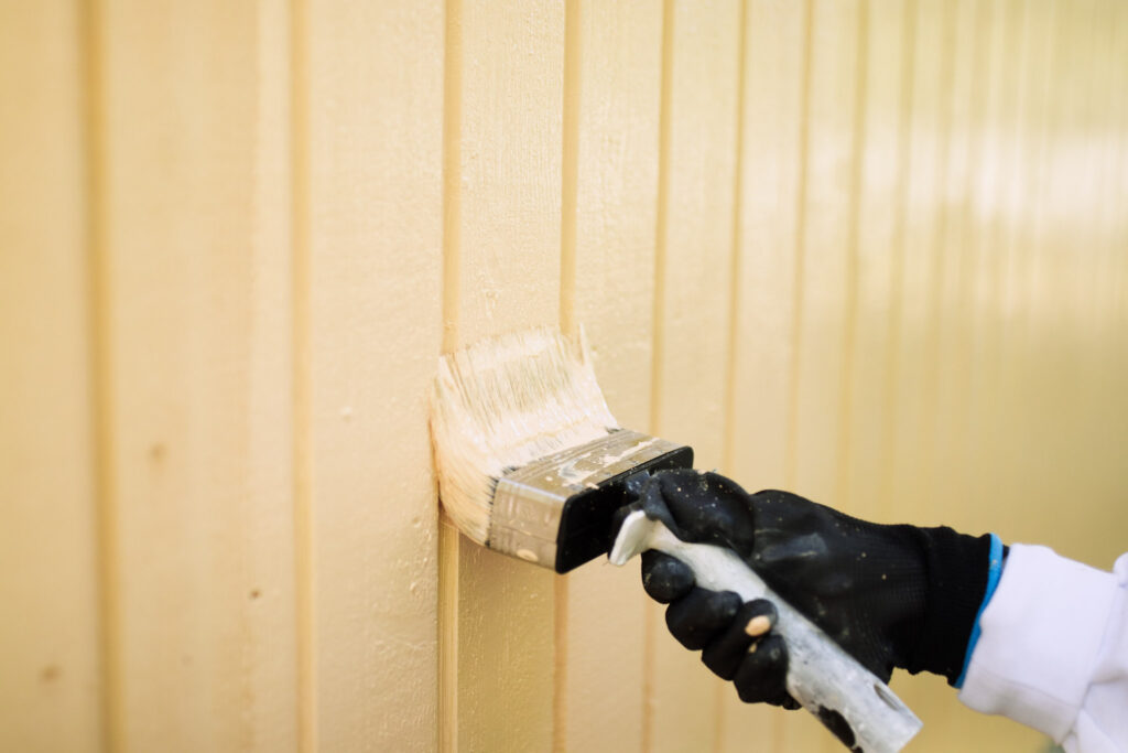 Maalari maalaa keltaista puuseinää aurinkoisella säällä kädessään hanska ja pensseli.