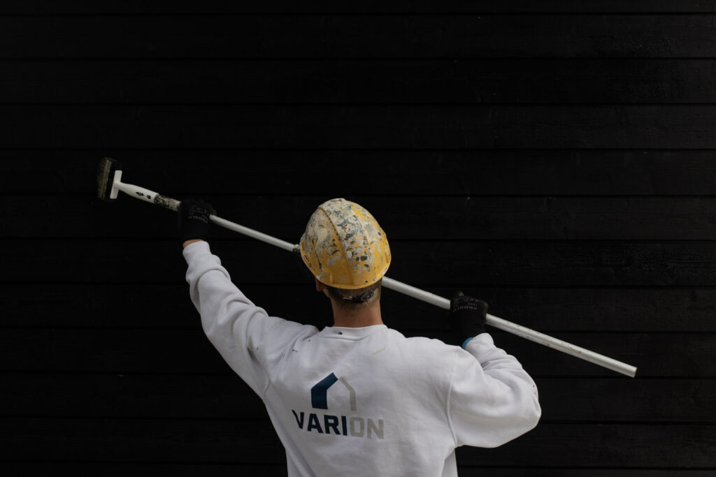 Maalari maalaa pensselillä ja jatkovarrella mustaa puuseinää