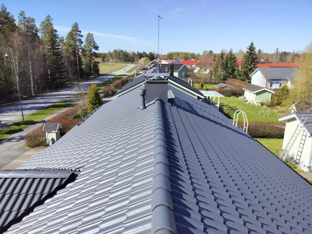 Musta tiilikatto ja savupiippu suomalaisella asutusalueella, ympärillä omakotitaloja, tie sekä nurmikkoa. Katolle on tehty tiilikaton pinnoitus.